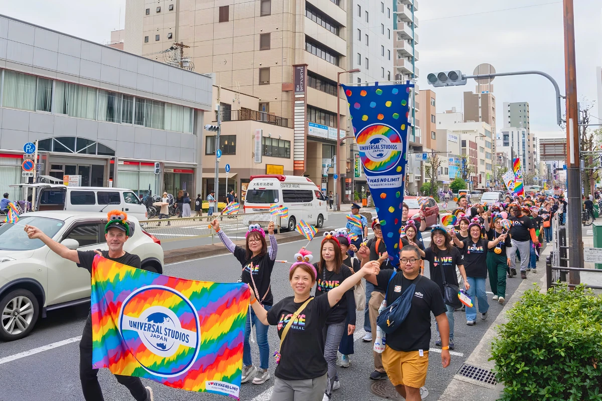 A large group of Universal Studios Japan staff and supporters march in a Pride parade, carrying rainbow flags and a colorful “Universal Studios Japan” banner along a city street.