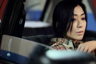 Hikaru Utada is sat in a car with the window down staring ahead with their hand raised near their face