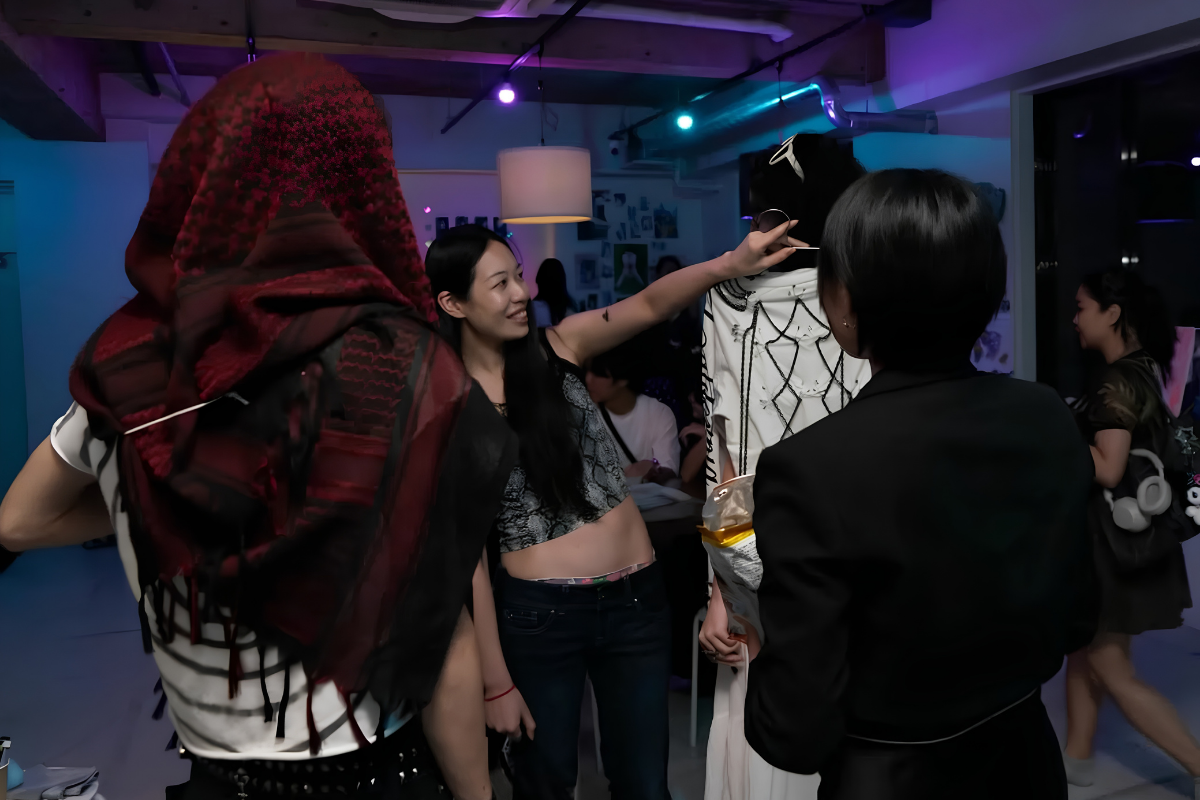 Leave the ROOM: Inside THEA's Queer-Inclusive Art Exhibition in Shinjuku 11 Two people, their backs to the viewer, examine art at a THEA exhibition. On the left, the visitor is wearing a dark red headscarf. In the center, a smiling artist explains their work