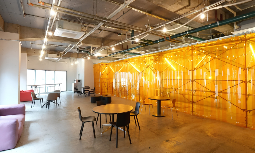 Interior of the WPÜ Shinjuku Gallery. Tables and chairs are sat at the center, while in the background and on the right large translucent plastic strips cover a developing exhibit