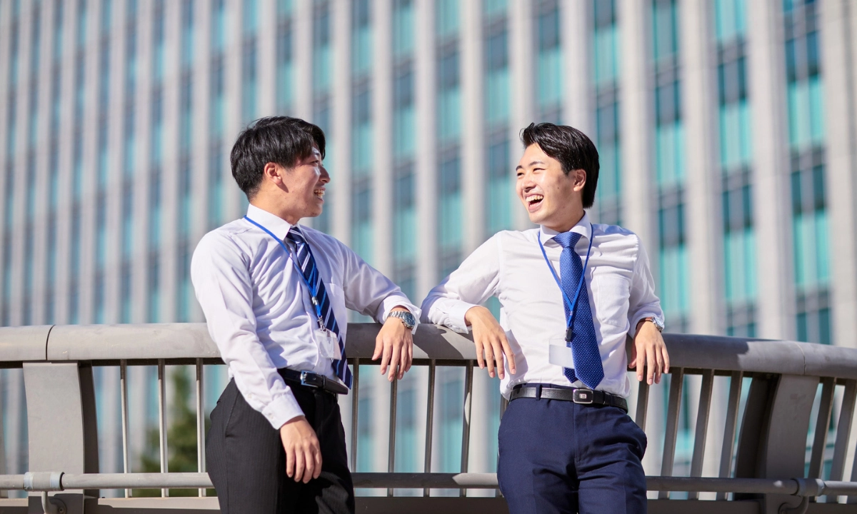 Two Japanese office workers in shirts and ties laugh together while talking outdoors beside a modern office building.