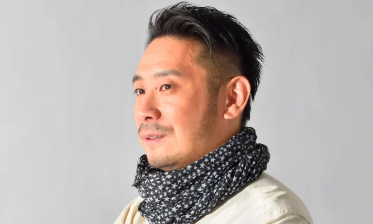 Portrait of a Japanese man with short dark hair and a trimmed beard, wearing a patterned scarf against a plain gray background.