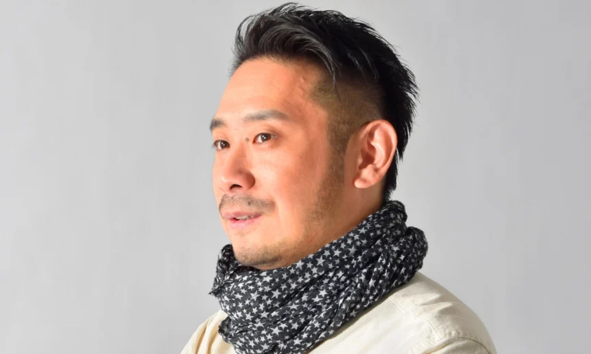 Portrait of a Japanese man with short dark hair and a trimmed beard, wearing a patterned scarf against a plain gray background.