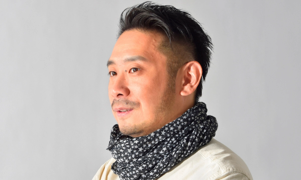 Portrait of a Japanese man with short dark hair and a trimmed beard, wearing a patterned scarf against a plain gray background.