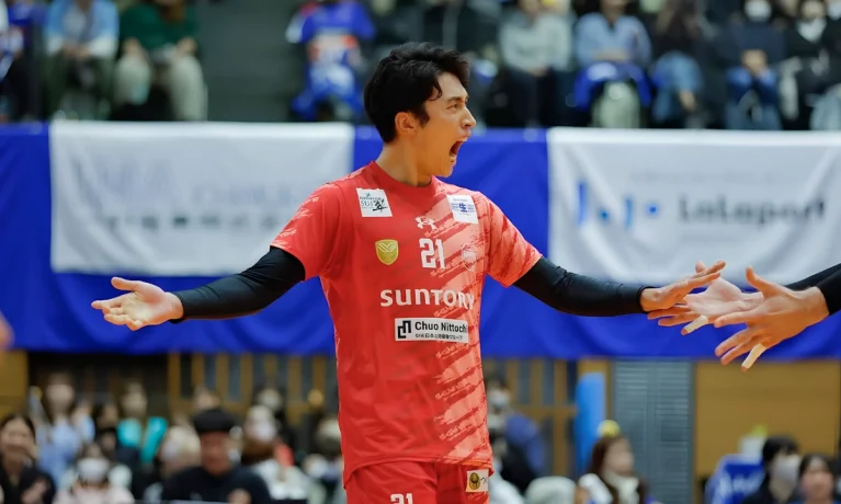 Male volleyball player in a red Suntory uniform celebrates on court with his arms outstretched